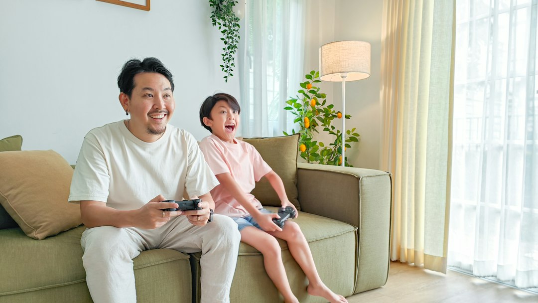 A father and child smile while playing a video game together on their living room couch.